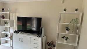 Una sala de estar con un televisor de pantalla plana sobre un mueble blanco, rodeada de estanterías blancas con plantas en macetas.