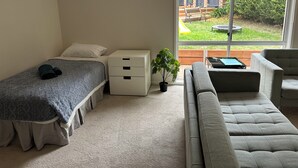 Un dormitorio con cama, mesita de noche, cómoda, ventana con vista a un jardín trasero y un sofá gris.
