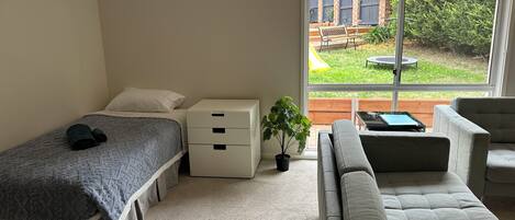 A bedroom with a bed, a nightstand, a dresser, a window with a view of a backyard, and a grey sofa.