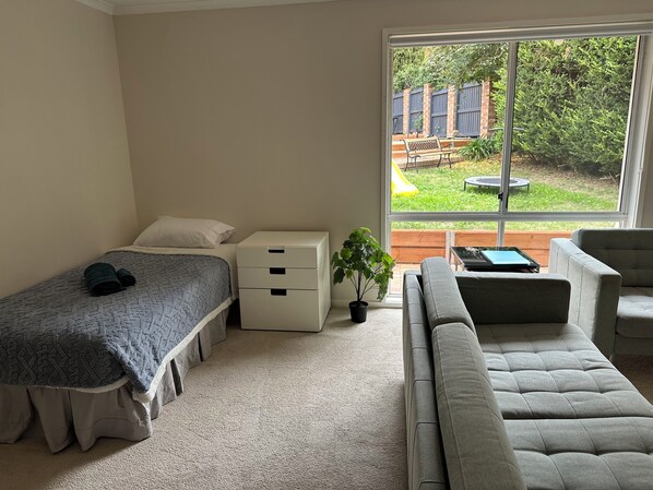 Een slaapkamer met een bed, een nachtkastje, een dressoir, een raam met uitzicht op een achtertuin en een grijze bank.