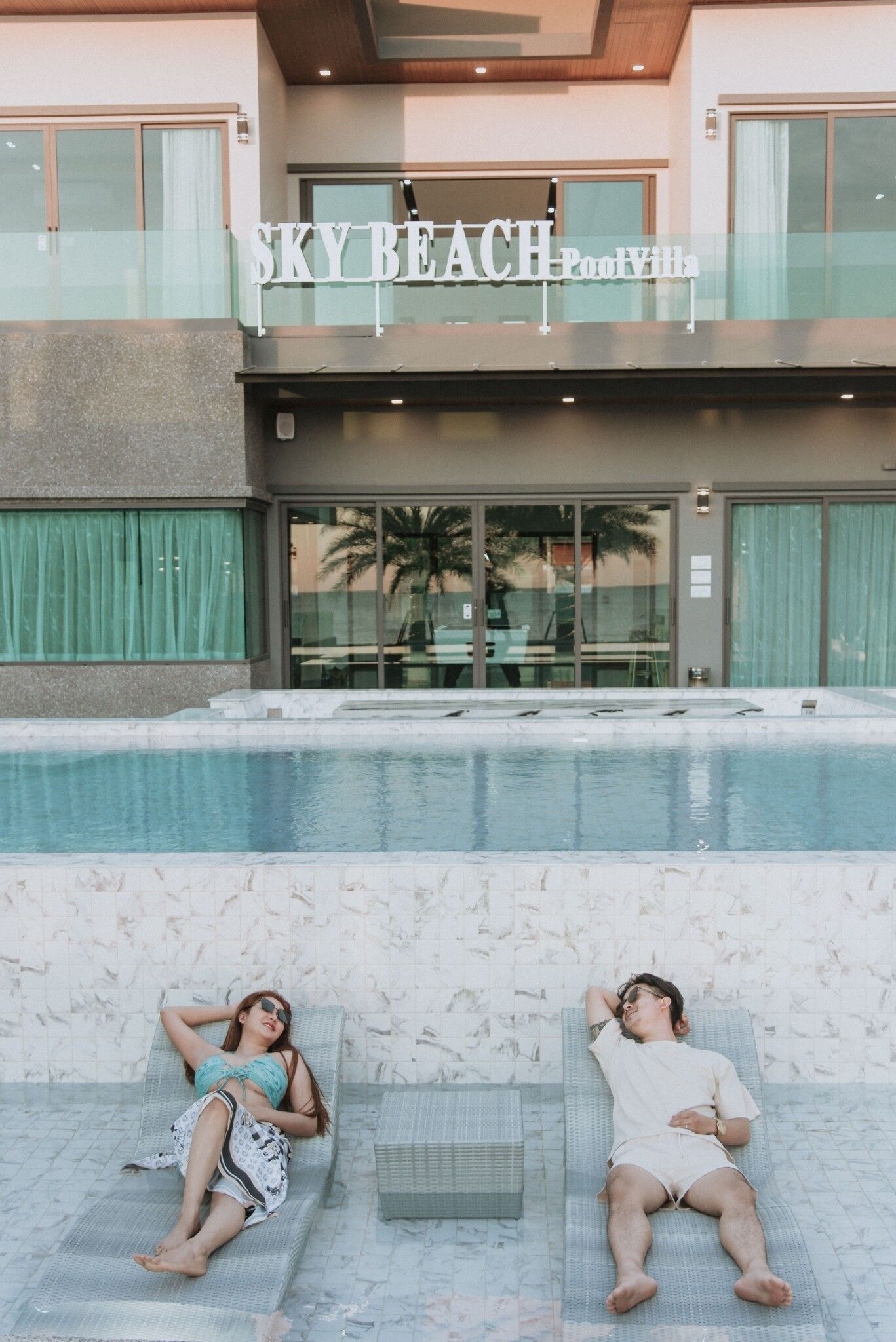 Un área de piscina con sillones de descanso, un edificio con un cartel que dice 'SKY BEACH Pool Villa' y una vista despejada de la piscina.