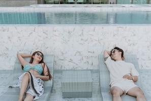 Two lounge chairs by a pool with a tiled wall and a small table in between.
