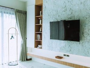 A modern living room with a TV mounted on a wall, a wooden shelf with decorative items, and a glass table.