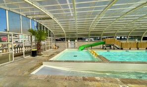 Piscina interna com escorregador, paredes de vidro e uma palmeira.