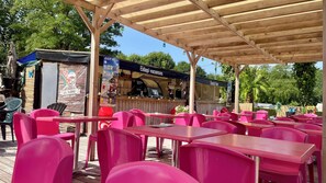 Ein Außenbereich mit Holztischen und -stühlen, ein überdachter Bereich mit einem Schild für 'Eldapi vacations' und eine Snackbar im Hintergrund.