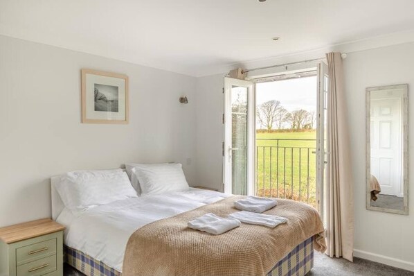 A bedroom with a bed, a nightstand, a framed picture, a door, and a view of a grassy area. - 4 bed lodge Newquay Cornwall Holiday Park sleeps 8 (Cornwall)