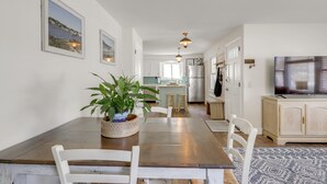 Uma área de refeições com mesa de madeira, cadeiras brancas, um vaso com planta e uma TV em um armário.
