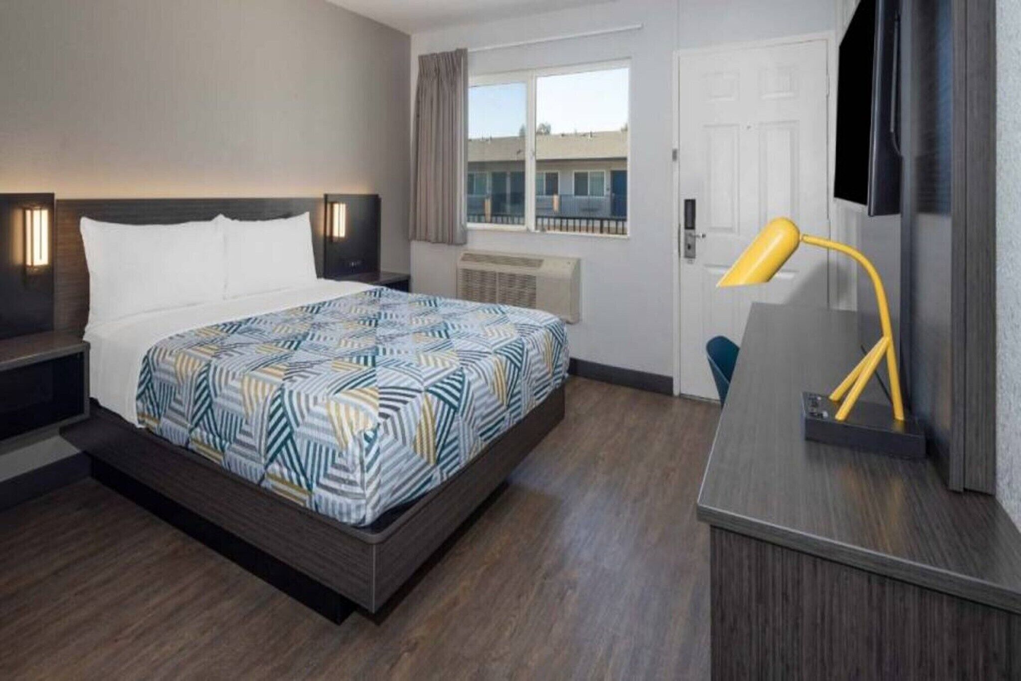 A hotel room with a bed, bedside tables, a desk, a lamp, a window, and an air conditioning unit.