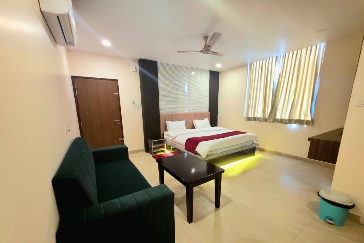 Una habitación de hotel con cama, sofá verde, una mesita, ventilador de techo y ventana con cortinas.