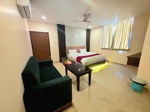Una habitación de hotel con cama, sofá verde, una mesita, ventilador de techo y ventana con cortinas.