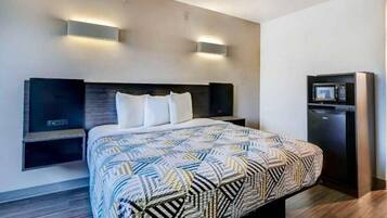 A modern hotel room with a large bed, bedside tables, a built-in TV, and a refrigerator.