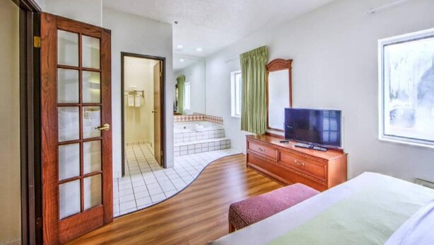 A hotel room with a wooden floor, a bed, a dresser, a television, and a bathroom visible through an open door. - 1 King Bed|Suite,Smk,Hot Tub (Dallas)