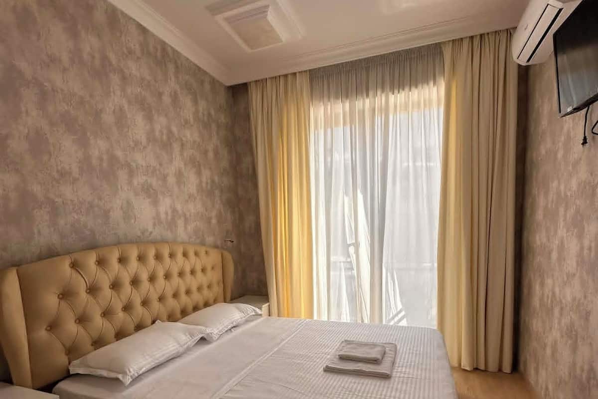 A bedroom with a tufted headboard, a bed with white linens, a bedside table, a flat-screen TV mounted on the wall, and a window with curtains.