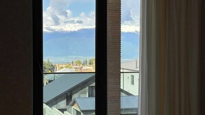 Un dormitorio con un gran ventanal corredero que da a un balcón con vistas a edificios y montañas.