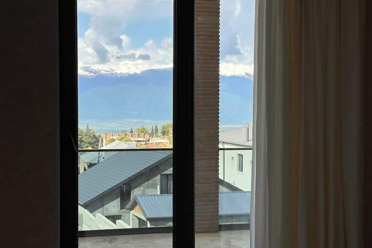 A bedroom with a large sliding glass door leading to a balcony with a view of buildings and mountains.