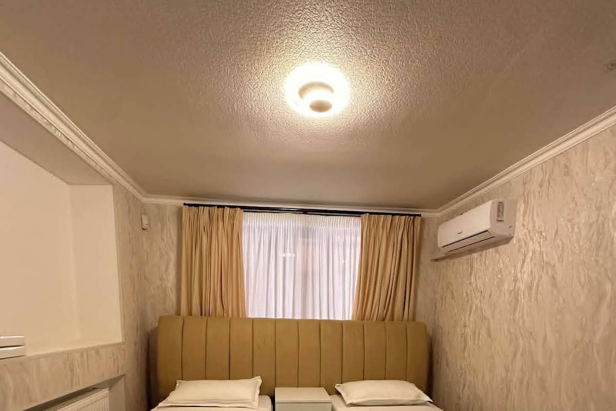 A room with two single beds, a nightstand, a window with curtains, an air conditioning unit, and a radiator.