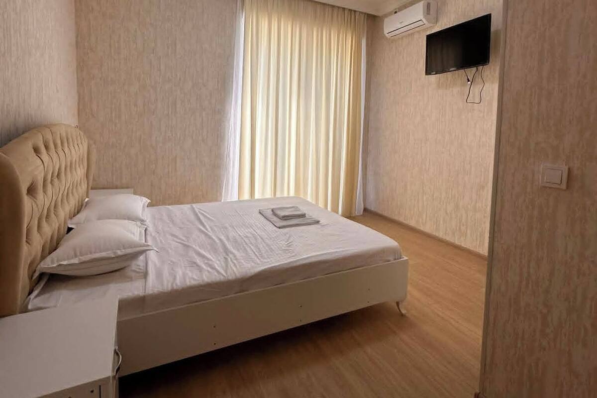 A bedroom with a bed, a bedside table, a flat-screen TV mounted on the wall, and a ceiling light.