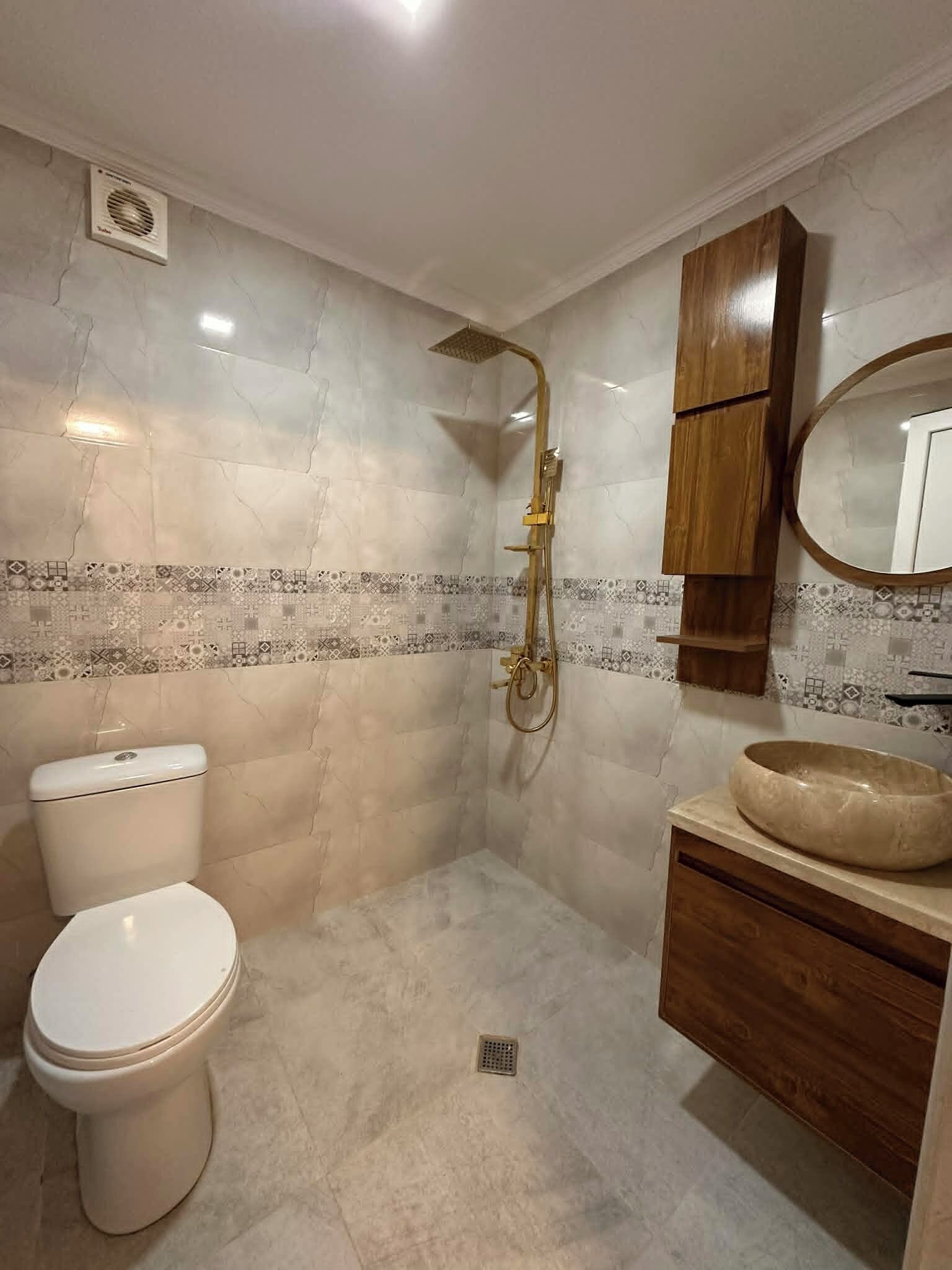A bathroom with a toilet, a shower with a golden showerhead, a wooden vanity with a round sink, and a mirror.