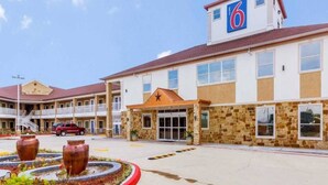 Un hôtel reconnaissable à son grand panneau indiquant le chiffre « 6 », une entrée centrale dotée d’un espace abrité et un parking où se trouve un véhicule.