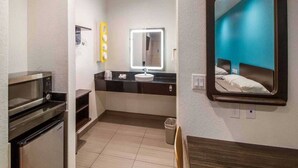 A compact hotel room with a kitchenette, a mirror with a light, and a bed with a headboard.