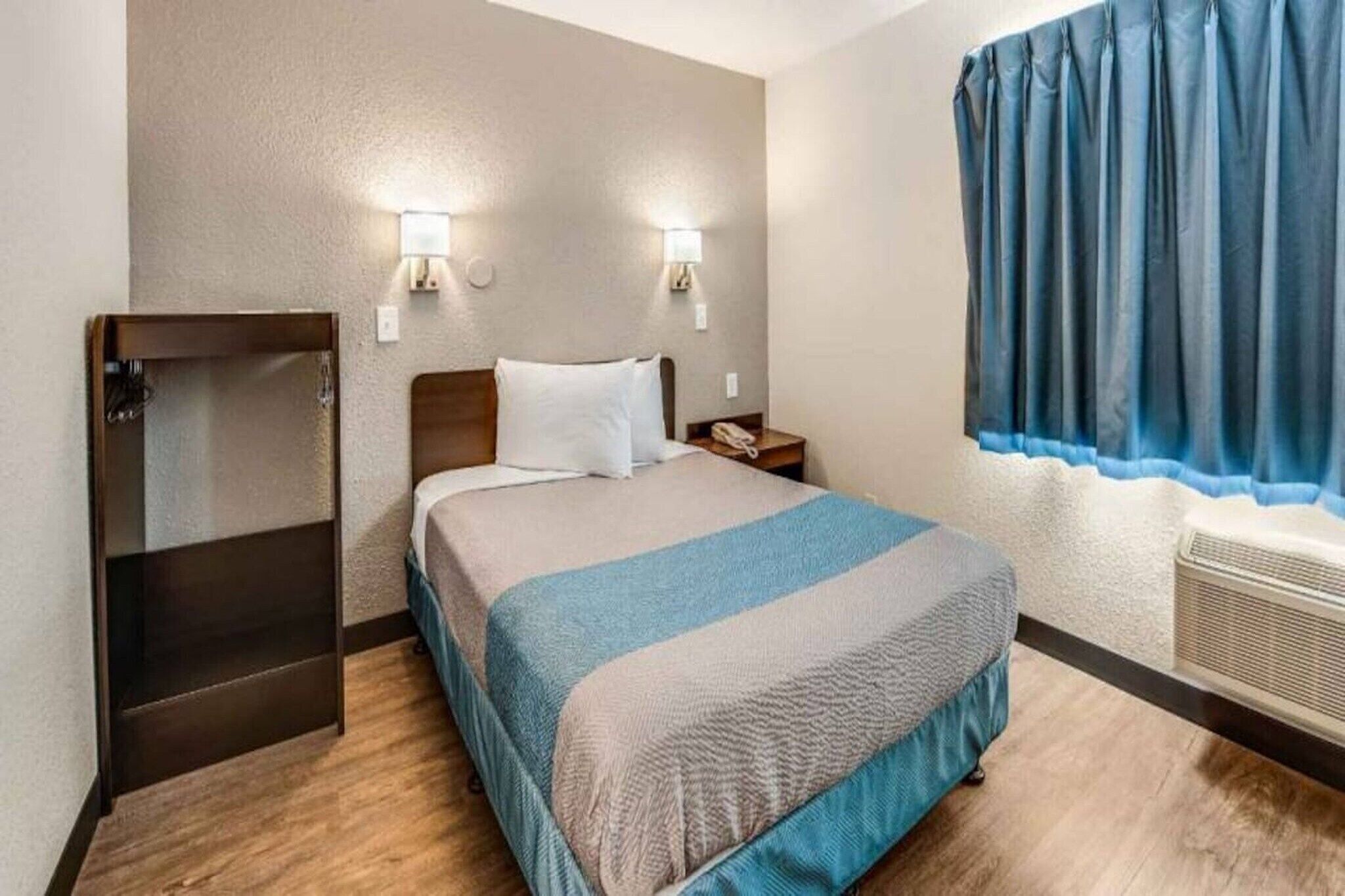 A hotel room with a bed, bedside lamps, a window with curtains, a heater, and a wardrobe.