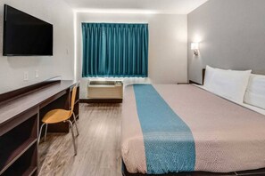 Hotellihuone, jossa on sänky, työpöytä tuolilla, taulutelevisio, ikkuna verhoilla ja ilmastointilaite.