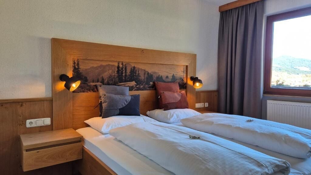 A double bed with two pillows, a nightstand, a headboard with a landscape painting, and a window with curtains.