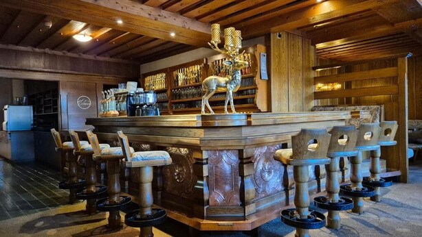 A bar area with a wooden ceiling, a deer sculpture, and a bar counter with stools. - Hotel Dahoam by Miha (Westendorf)