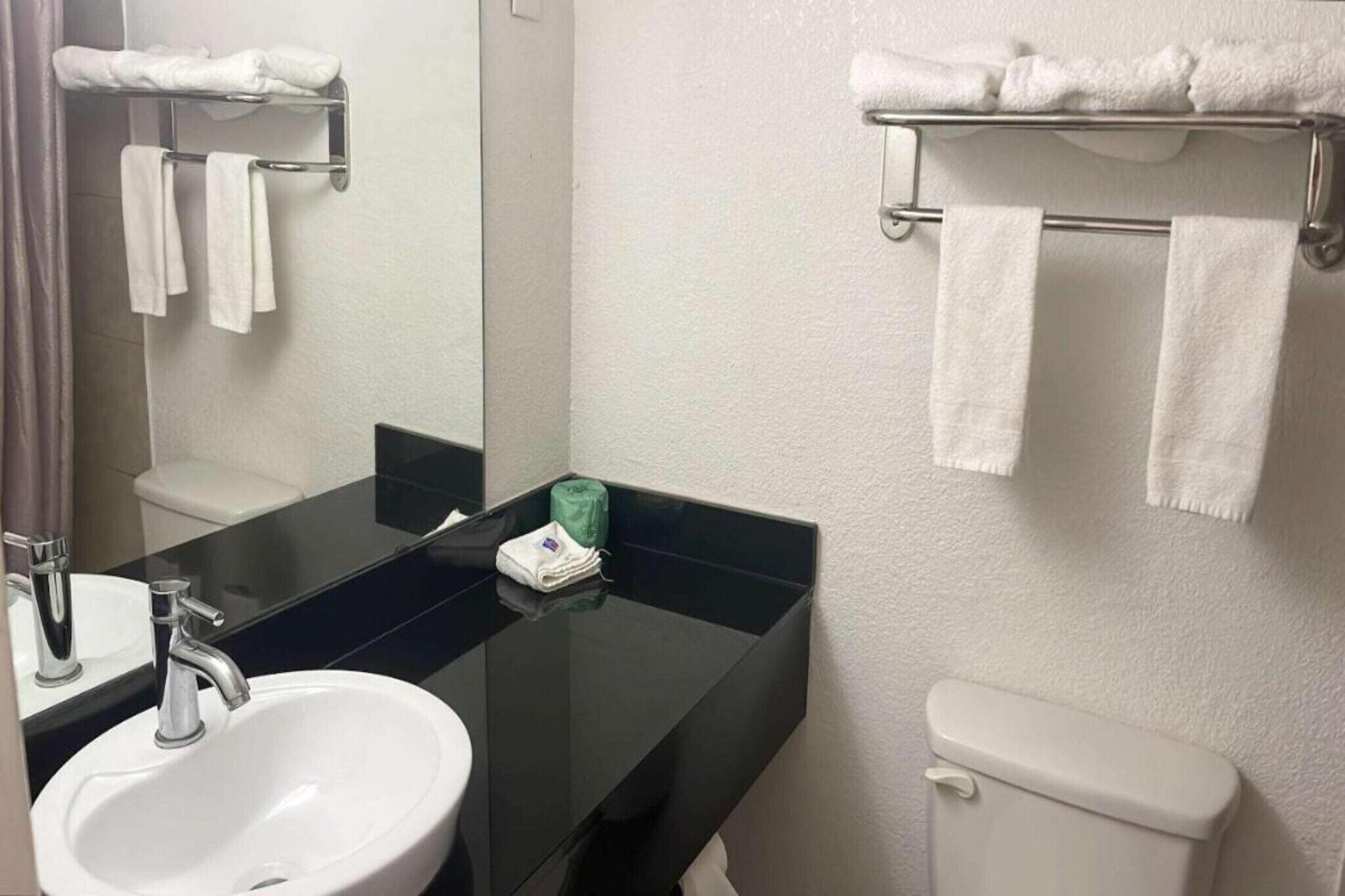 A bathroom with a black countertop, white sink, and toilet. There are white towels on a rack and a green bag on the countertop.