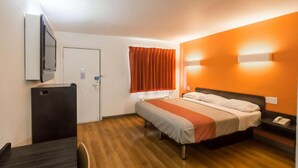 A hotel room with a bed, a desk, a chair, a TV, a window with curtains, and a wall-mounted light fixture.