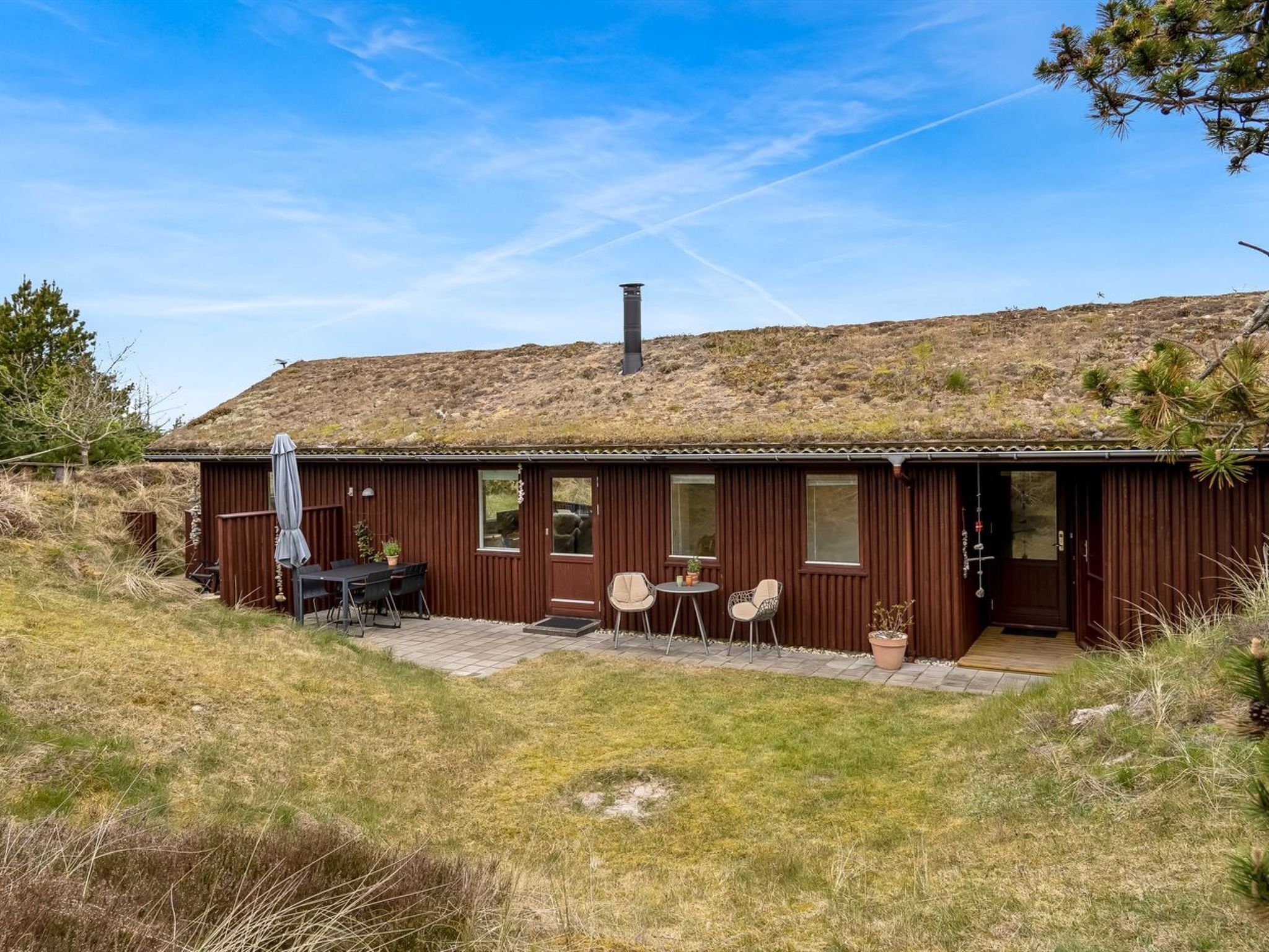 Une cabane rustique avec un toit plat, une cheminée, un patio avec des meubles d’extérieur et une vue sur une colline en arrière-plan.