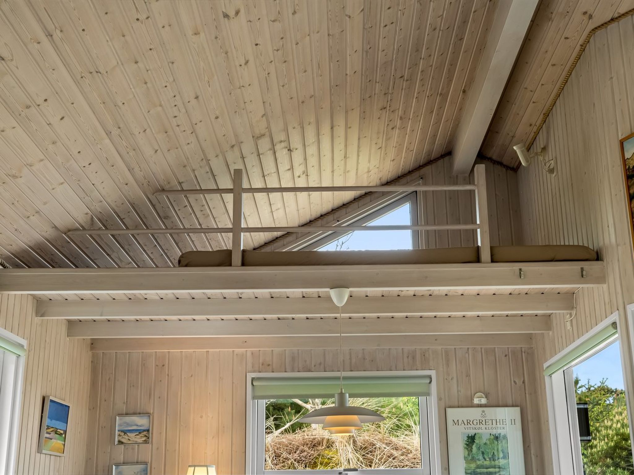 A wooden interior with a sloped ceiling, a window with a view of greenery, and a framed picture on the wall.