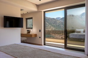 Ein modernes Schlafzimmer mit einem großen Bett, einem an der Wand montierten Fernseher, einem Schreibtisch mit Stuhl und einer Glastür, die auf einen Balkon mit Bergblick führt.