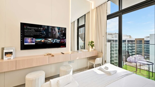 Una camera d'albergo moderna con una grande TV, un balcone con vista sulla città, una scrivania con un portatile e un letto con asciugamani.
