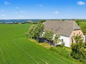 Une maison avec un toit en pente, entourée d’un grand champ verdoyant et avec une étendue d’eau en arrière-plan.