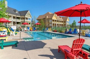 Ein Poolbereich mit bunten Adirondack-Stühlen und Sonnenschirmen, ein Pool mit Sprungbrett und ein Gebäude mit Balkon im Hintergrund.