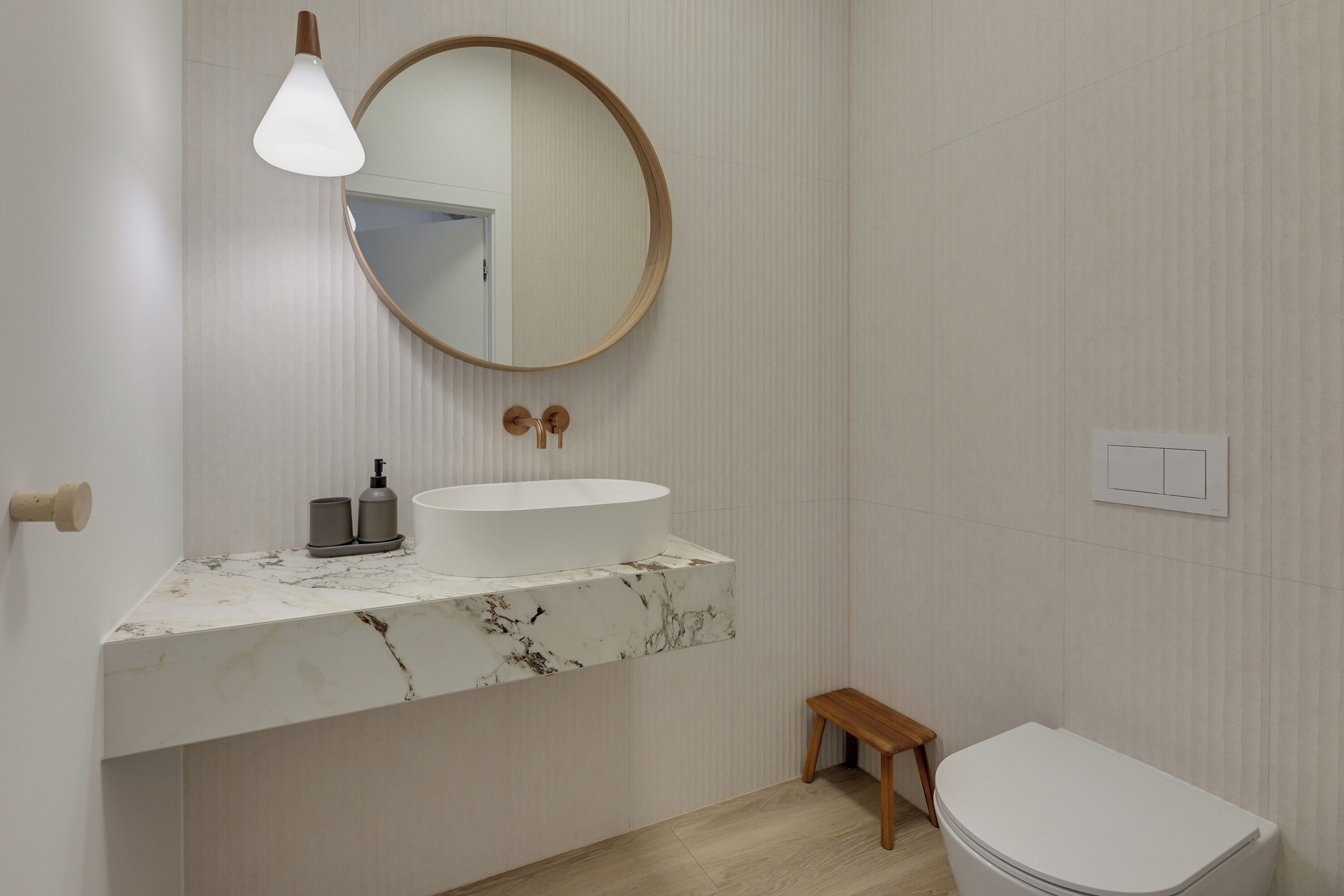 Ein Badezimmer mit Marmorwaschbecken, einem runden Spiegel, einem kleinen Holzschemel und einer weißen Toilette.