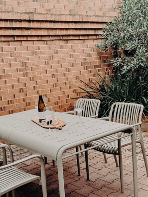 Une terrasse extérieure avec un mur en briques, une table en métal, des chaises et une bouteille de vin.