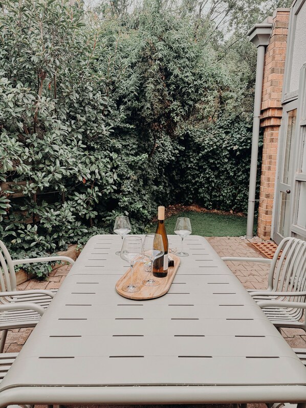 An outdoor patio with a table set for a meal, surrounded by greenery and a brick wall.