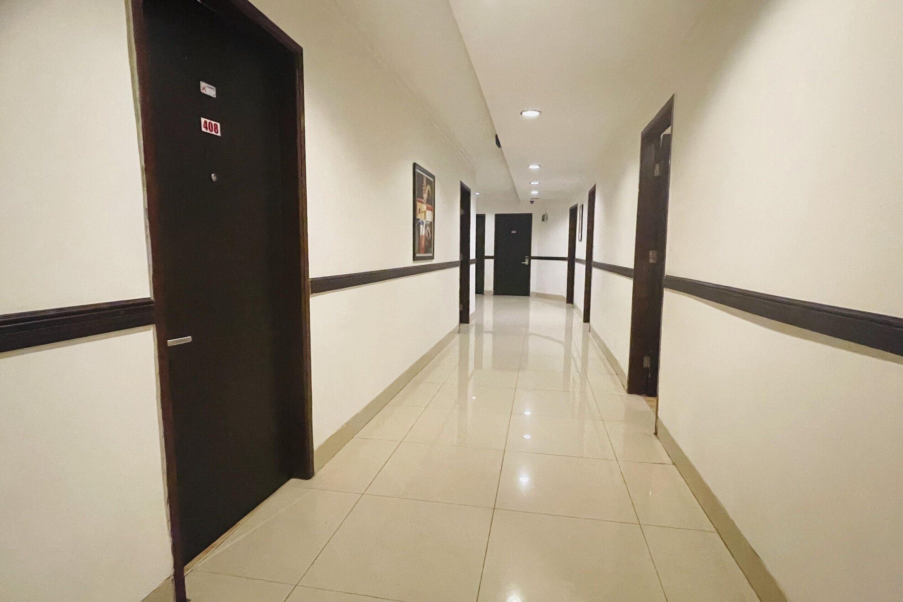 A hotel hallway with a dark wooden door on the left, a white tiled floor, and a series of doors along the corridor.