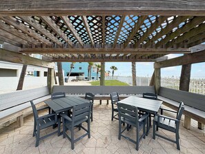 A covered outdoor seating area with wooden benches and tables, a wooden pergola overhead, and a view of a building and beach in the background. - Ocean House 2701 Beachfront (Gulf Shores)