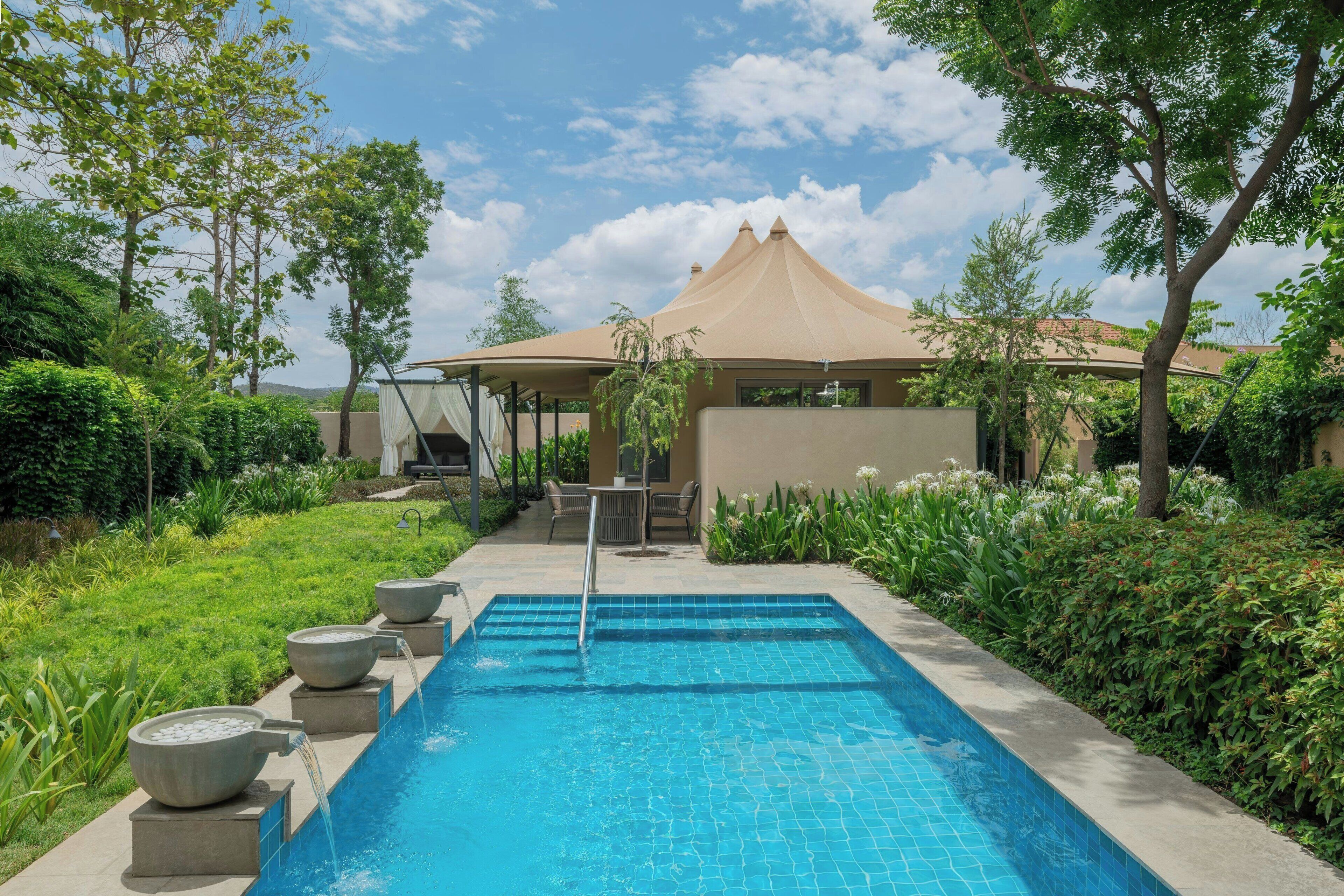 A swimming pool with a tiled edge, surrounded by greenery and a building with a conical roof.