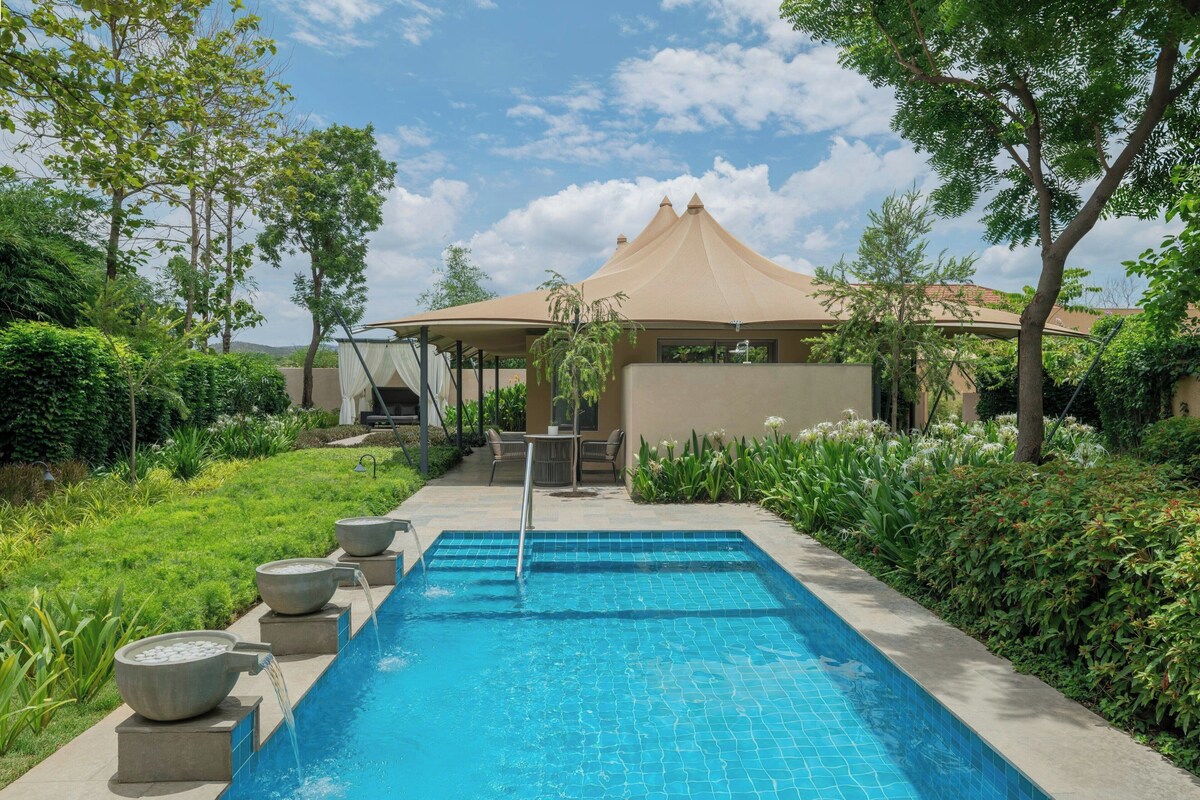 A swimming pool with a tiled edge, surrounded by greenery and a building with a conical roof.