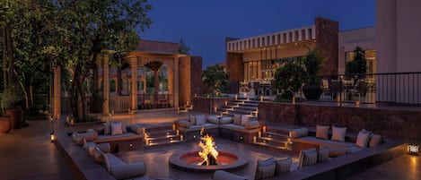 Un espacio exterior con zona de estar al aire libre, una chimenea, asientos acolchados y una escalera que lleva a un nivel superior.