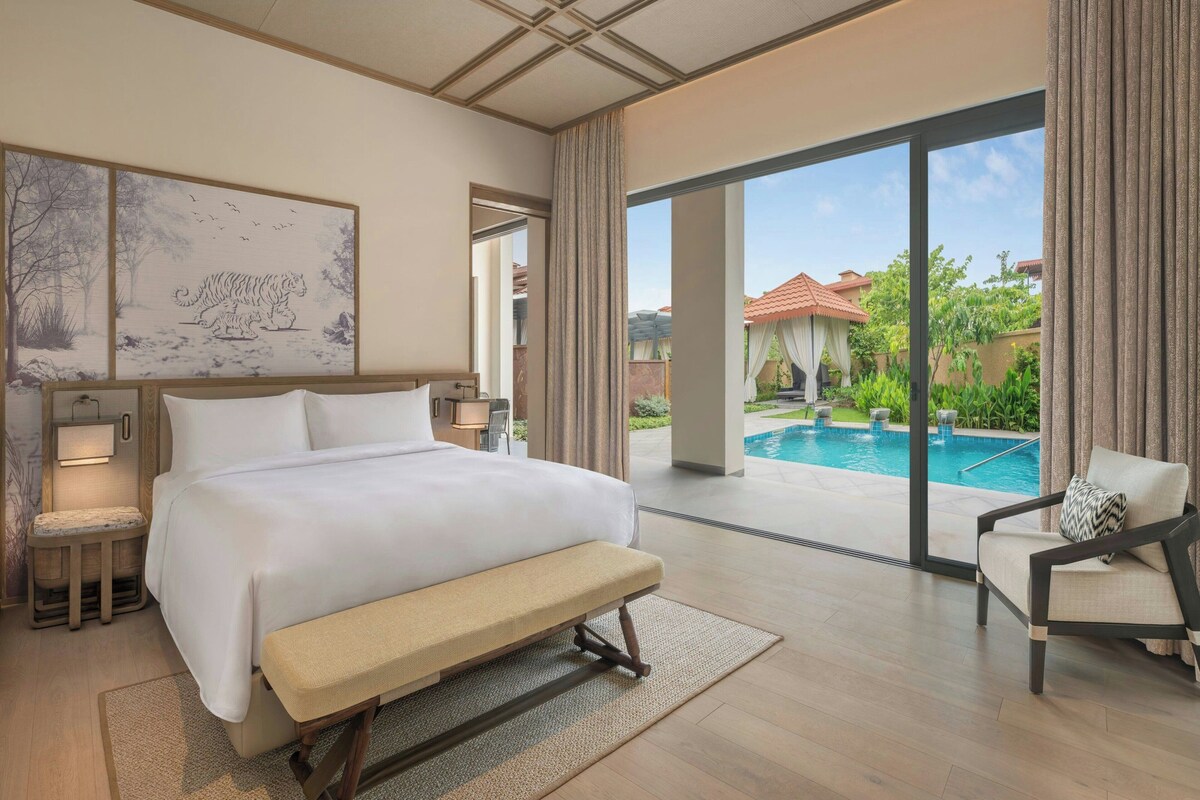 A hotel room with a large bed, a bench, a desk, a chair, and a view of a swimming pool.