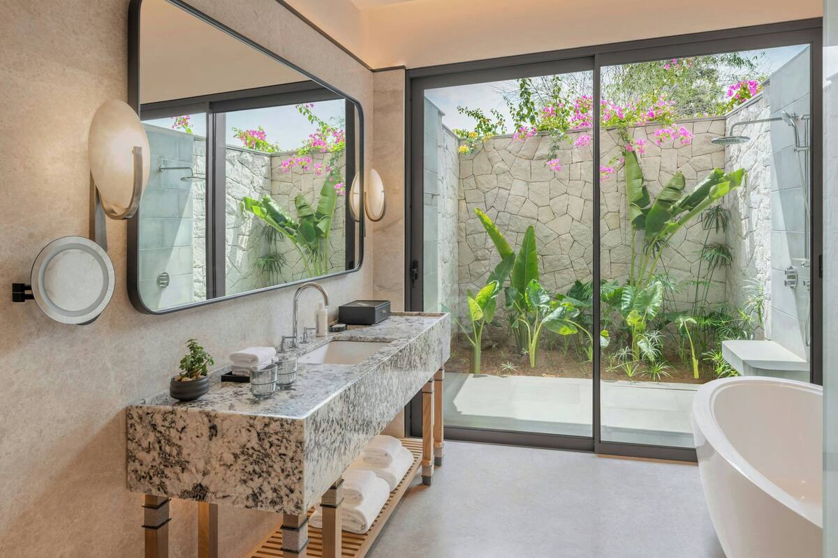 A modern bathroom with a large mirror, a granite countertop, a bathtub, and a view of a garden through a glass door.