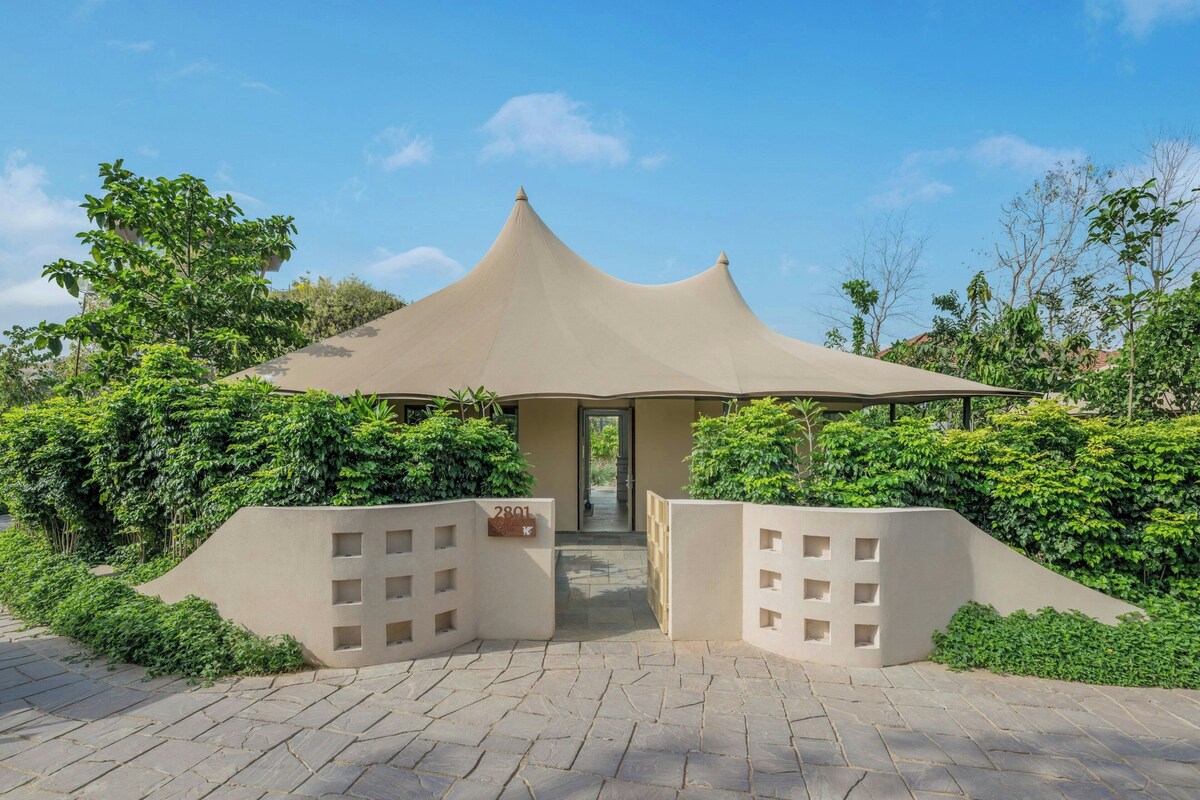 A building with a unique conical roof structure, surrounded by lush greenery and a paved entrance area.