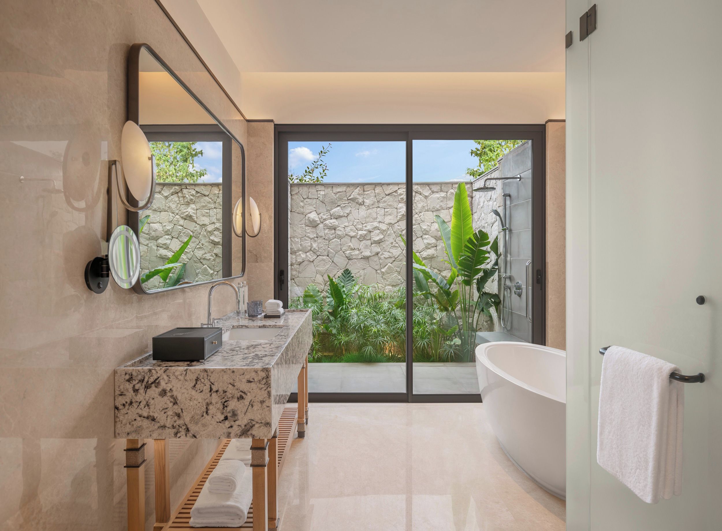 A modern bathroom with a large bathtub, a double vanity with a marble countertop, a stone wall, and a view of greenery.