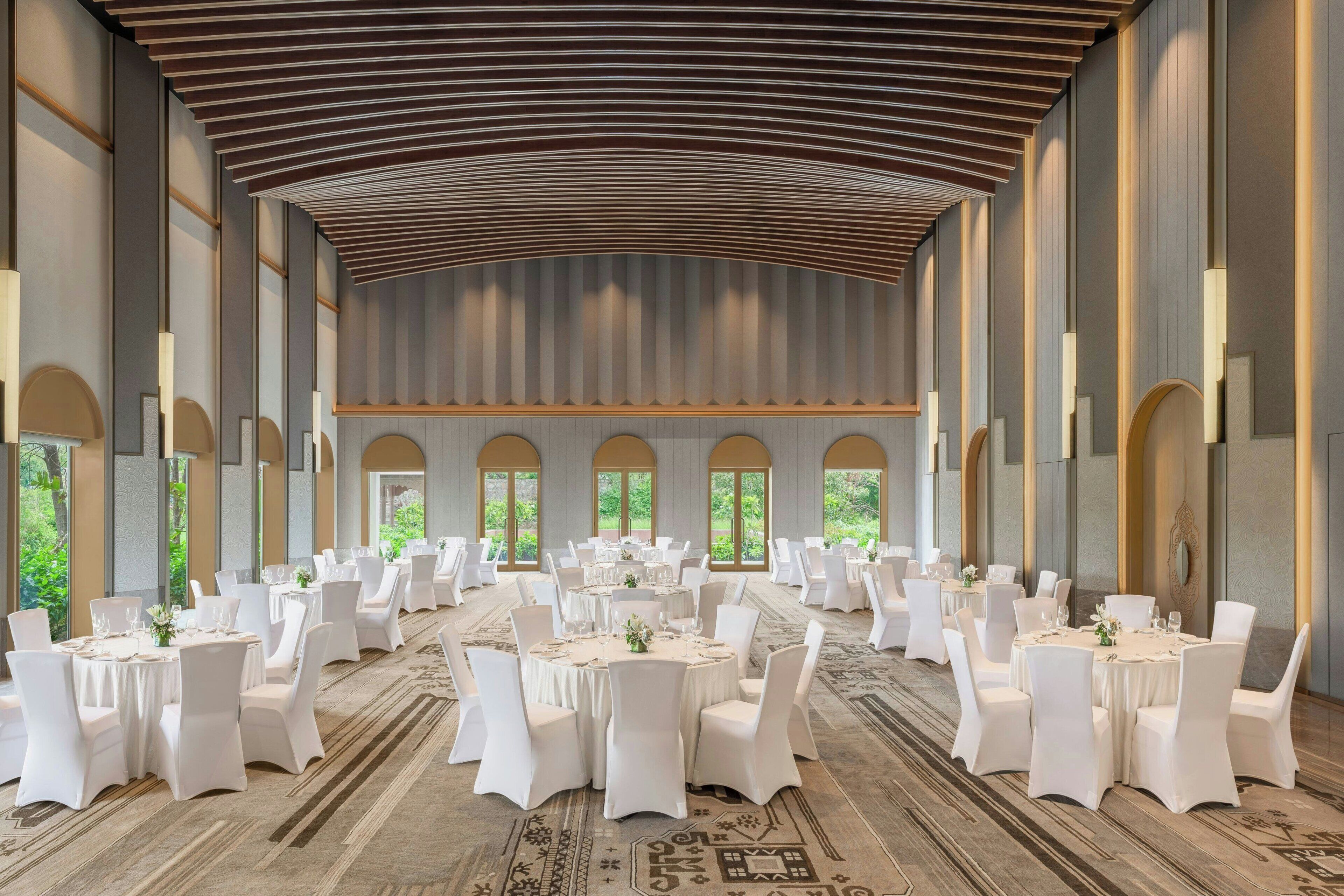 Une salle de banquet spacieuse, dotée de nombreuses tables rondes, de chaises blanches et d’un plafond haut avec des lambris en bois.