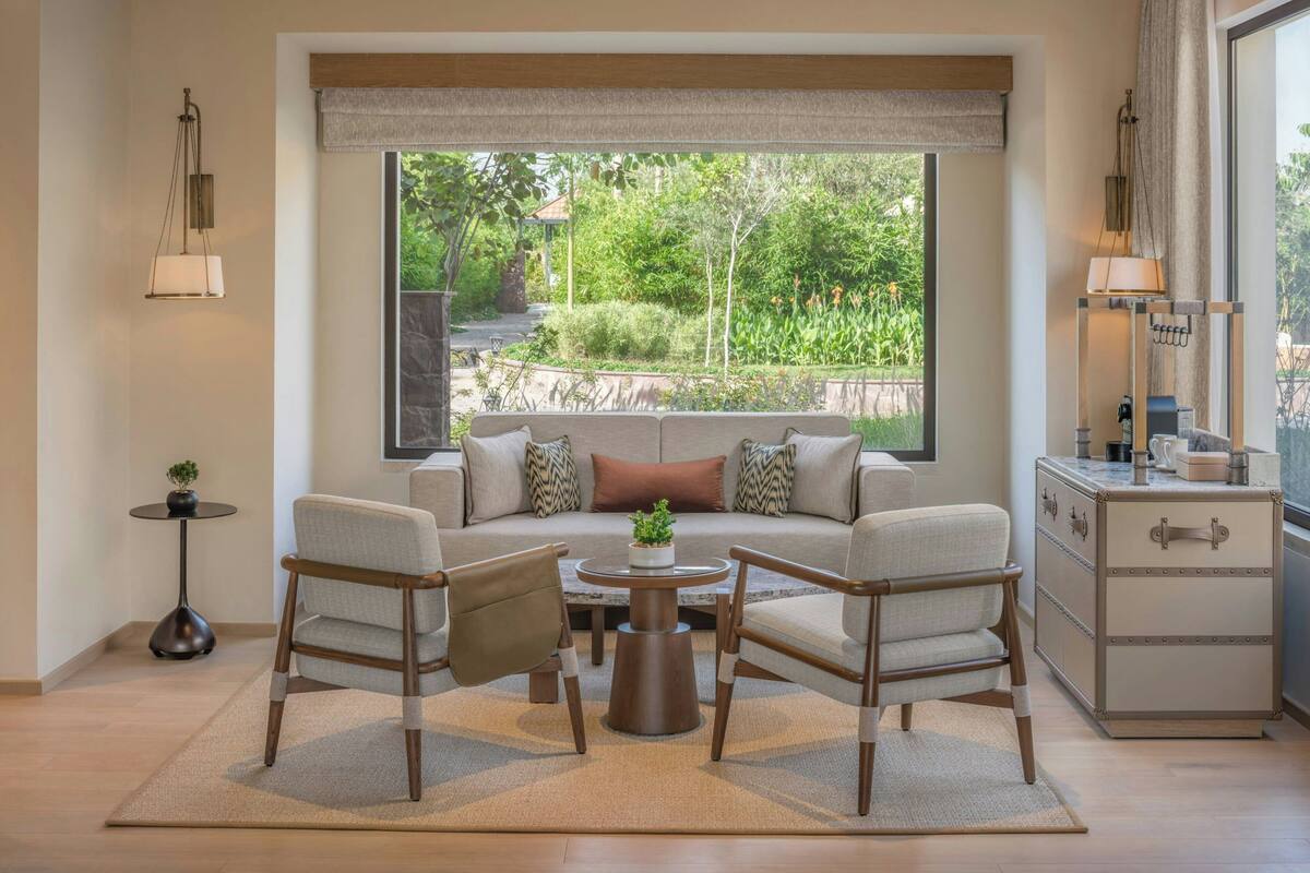 A living room with a sofa, two armchairs, a coffee table, a sideboard, and a large window with a view of greenery.
